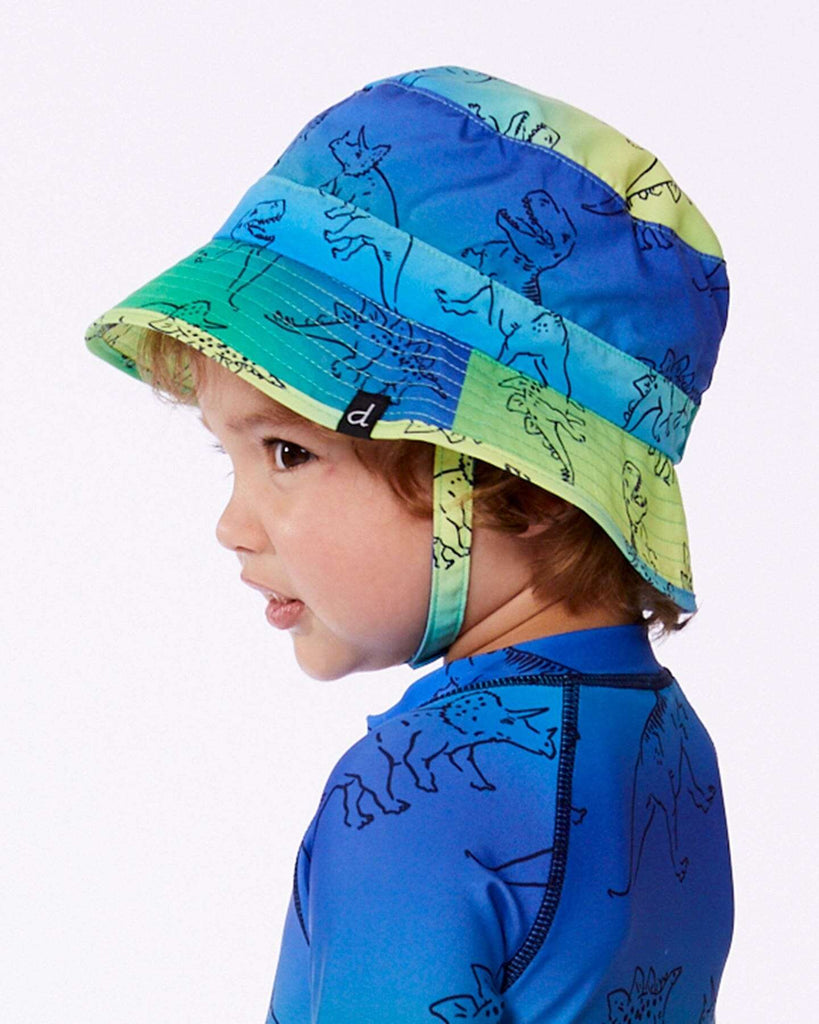 Little boy wearing blue dinosaur-patterned sun hat and matching swim shirt facing left on white background