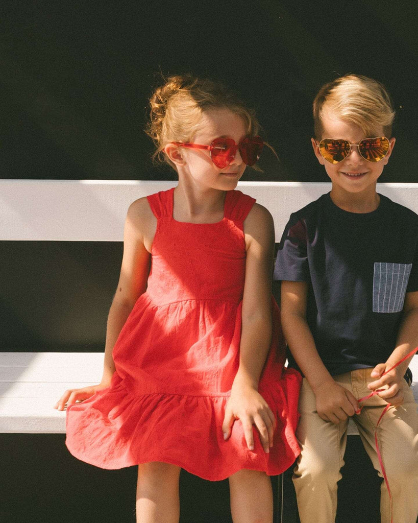 Girl wearing red multi-tiered eyelet dress with wide straps and square neckline sitting on a bench next to boy in sunglasses