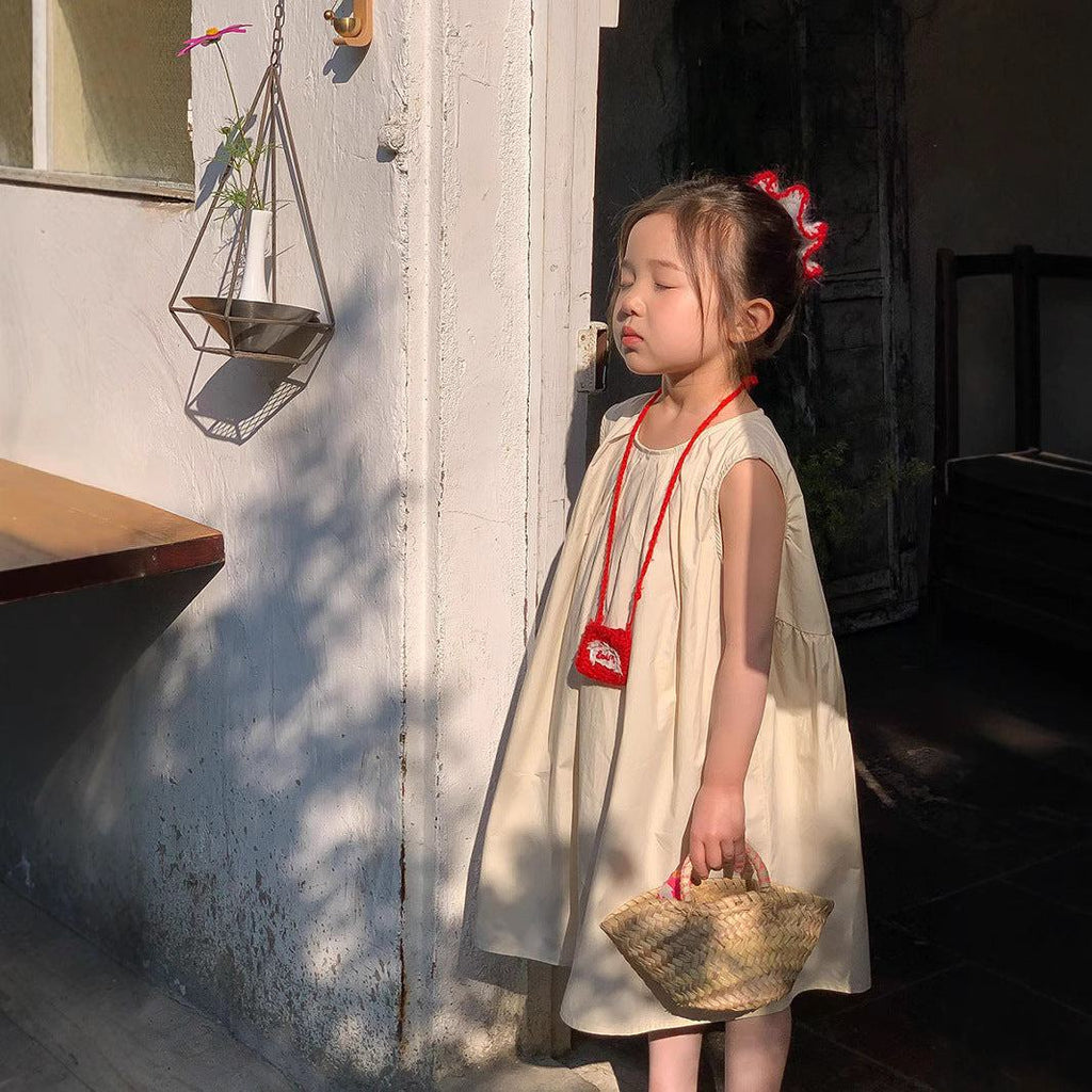 Petite fille portant une robe enfant douce et élégante sans manches en coton couleur abricot, tenant un petit panier en osier.