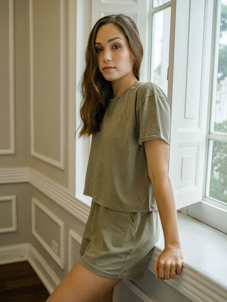 Woman wearing Creamfields Organic Cotton soft olive green pajama shorts and matching top by a window in a cozy room