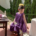 Fille portant une robe enfant douce sans manches de couleur violette avec jupe évasée en coton, assise dans un jardin au soleil
