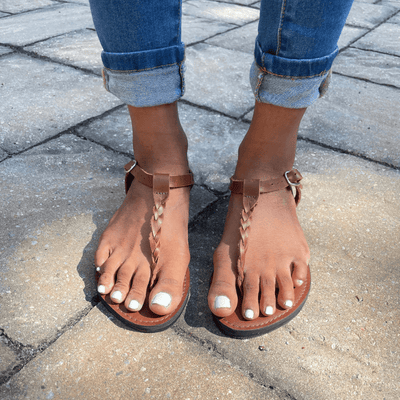Brown Bonita Roman Style Sandal with braided leather straps and ankle buckle worn with rolled-up jeans on stone pavement