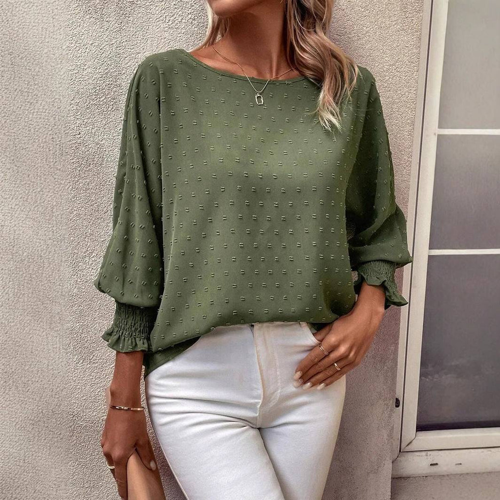 Woman wearing green round neck lantern sleeve polyester blouse with smocking detail and white pants standing by a wall and window