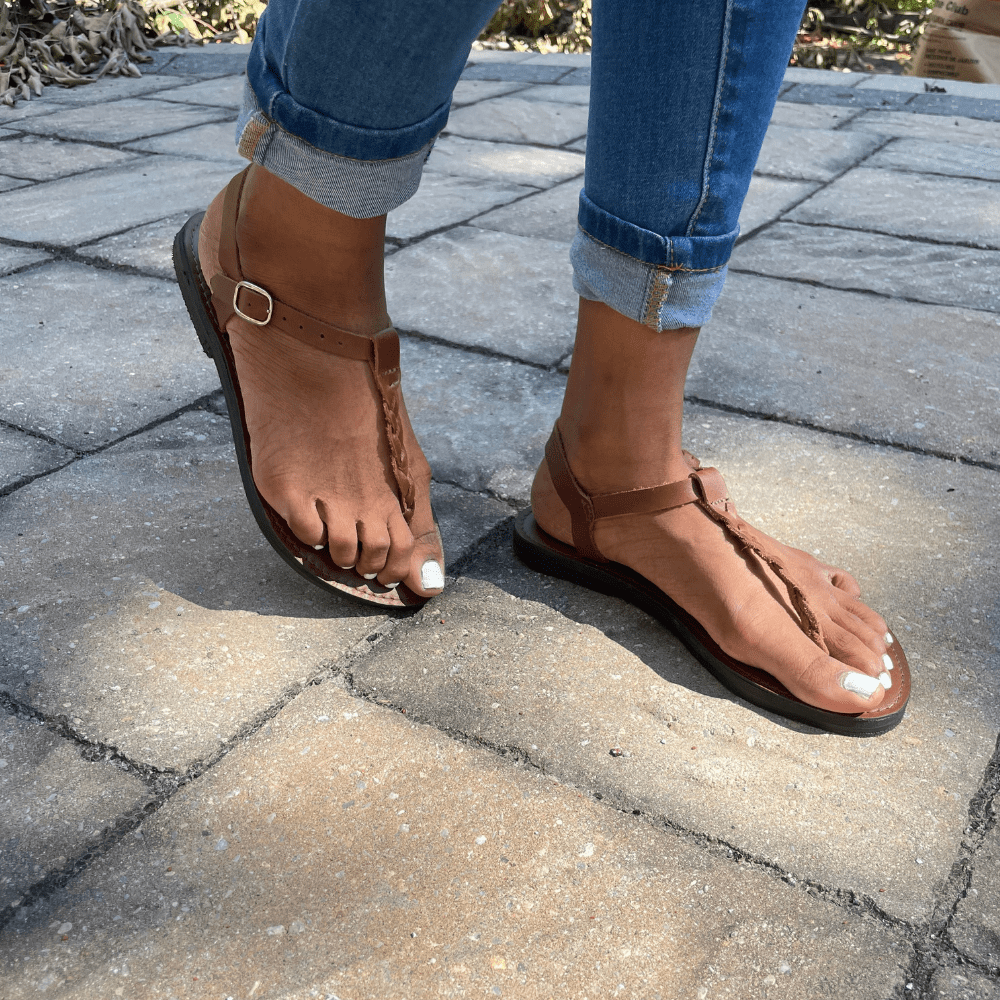 Brown Bonita Roman Style Sandal with ankle strap and cushioned sole worn with rolled-up jeans on stone pavement outdoors