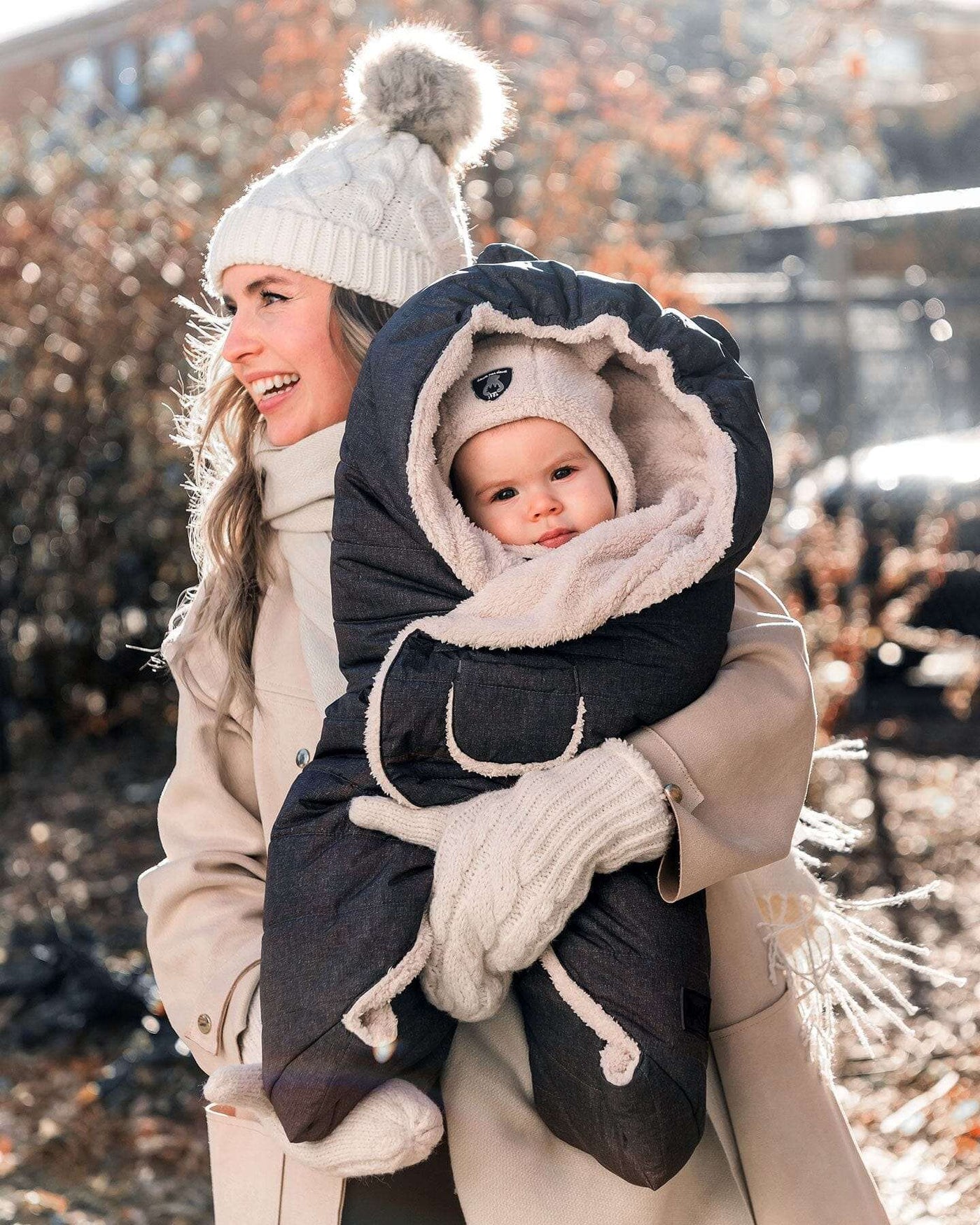 Baby wrapped in Snuggly Wrap Black for car seat and stroller with plush lining and animal ears, held by woman outdoors in winter clothing