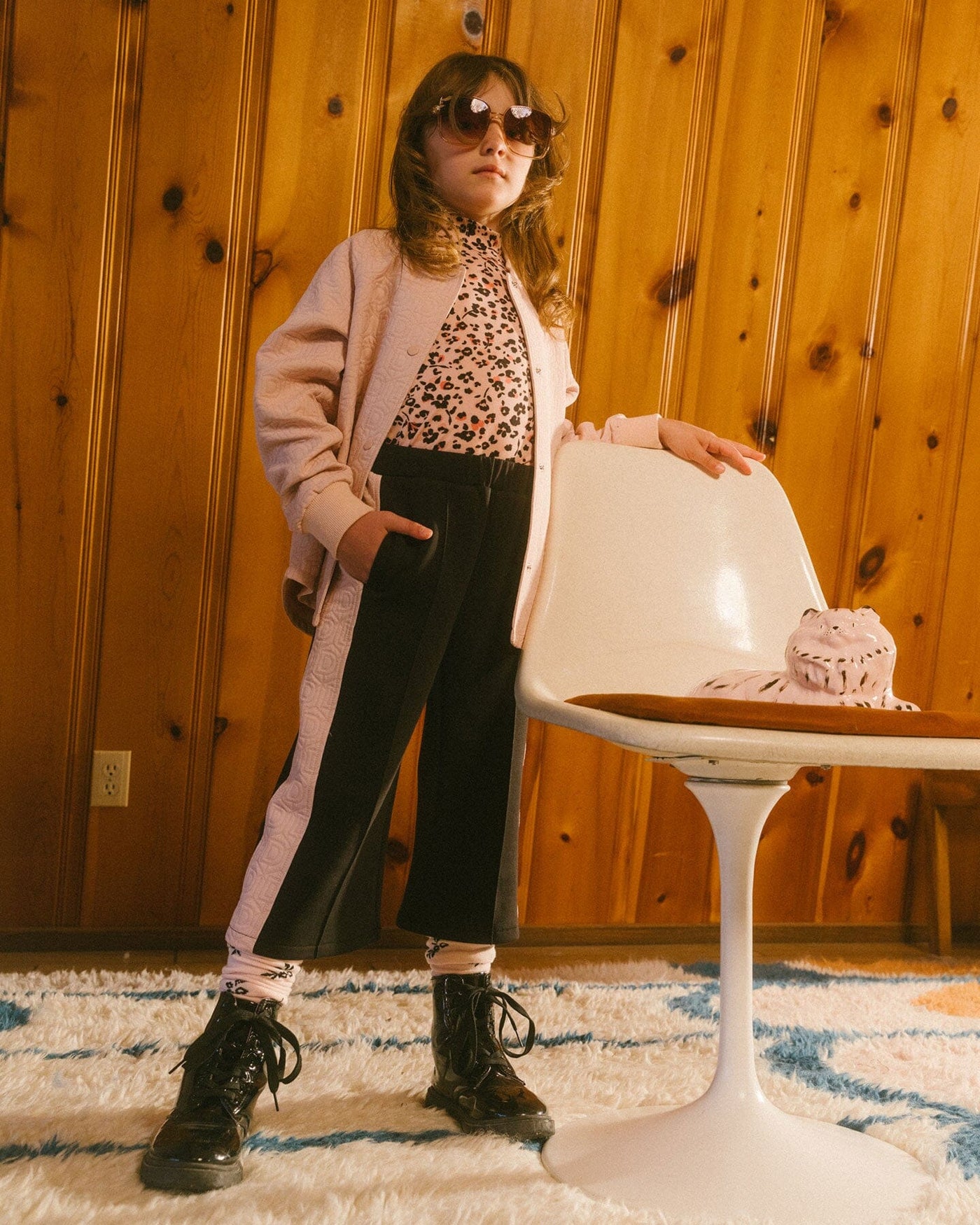 Child wearing black cropped wide leg pants with contrast pink side panels, paired with pink jacket and leopard print top, standing indoors against wood-paneled wall.