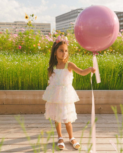 Little girl wearing a tiered printed mesh dress with butterfly appliques holding a pink balloon outdoors
