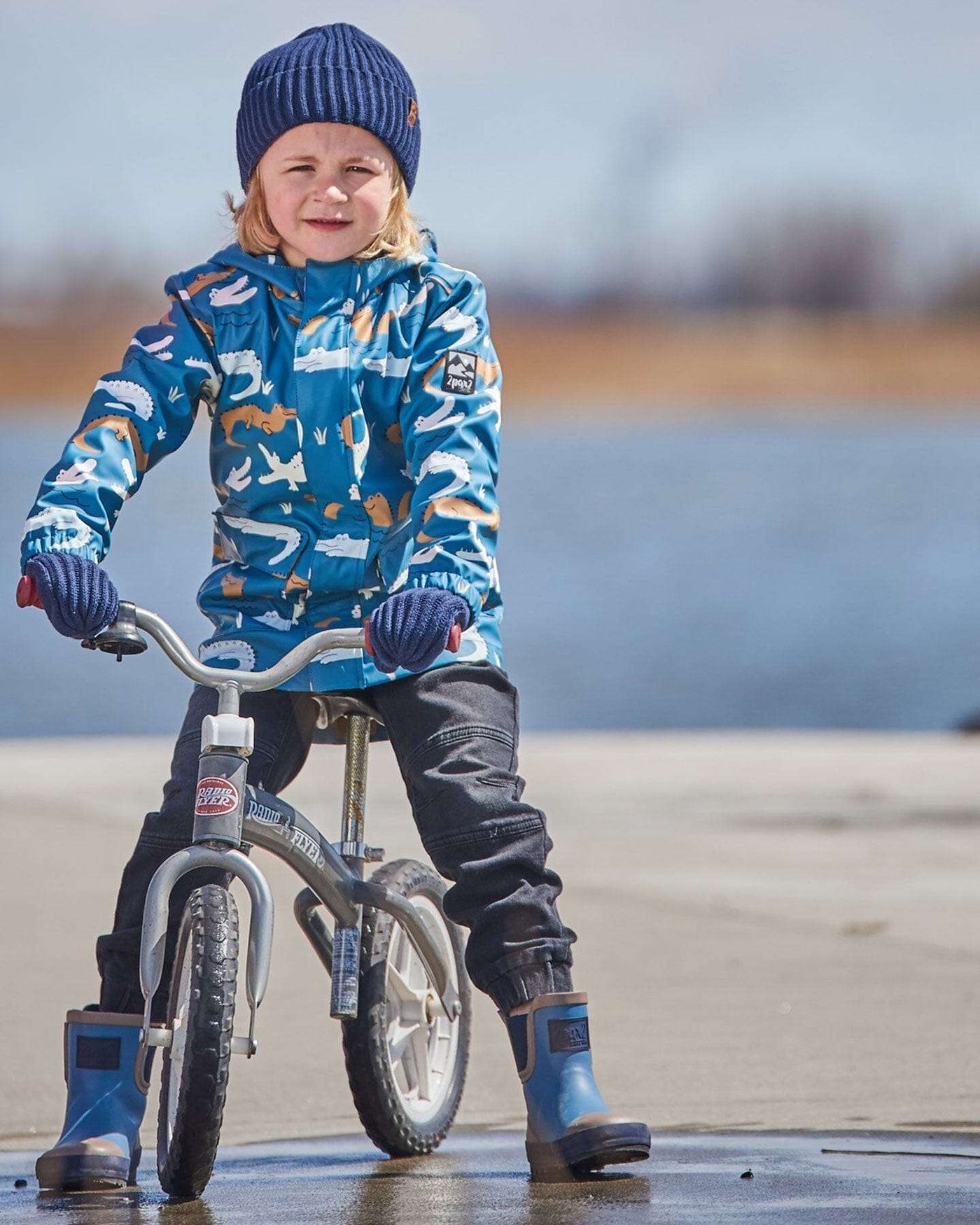 Child wearing a blue raincoat with color-changing crocodile print riding a bike by water.