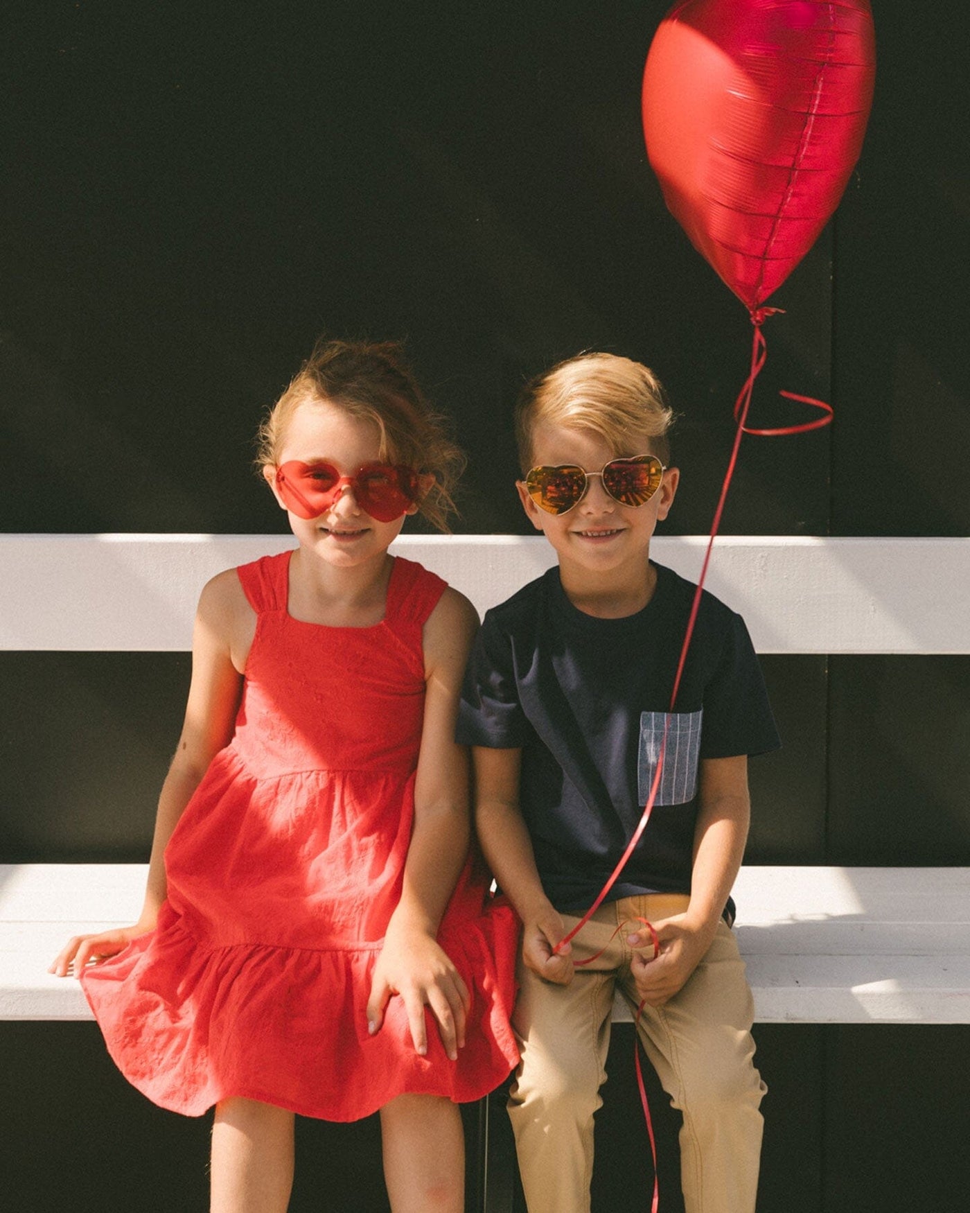 Children sitting on a bench wearing light golden beige stretch twill pants and colorful outfits with sunglasses and a red balloon