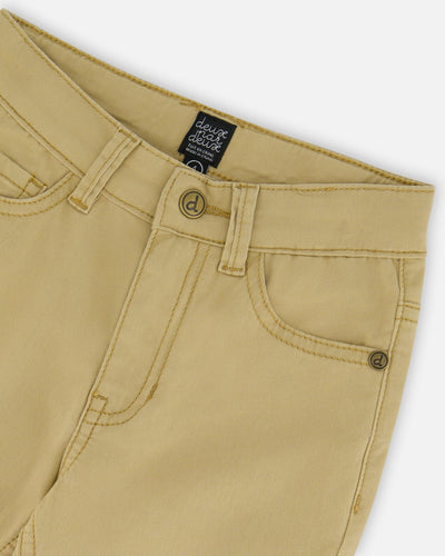 Light golden beige stretch twill pants with fixed waistband, zipper and button closure, front scoop pockets, and logo hardware.