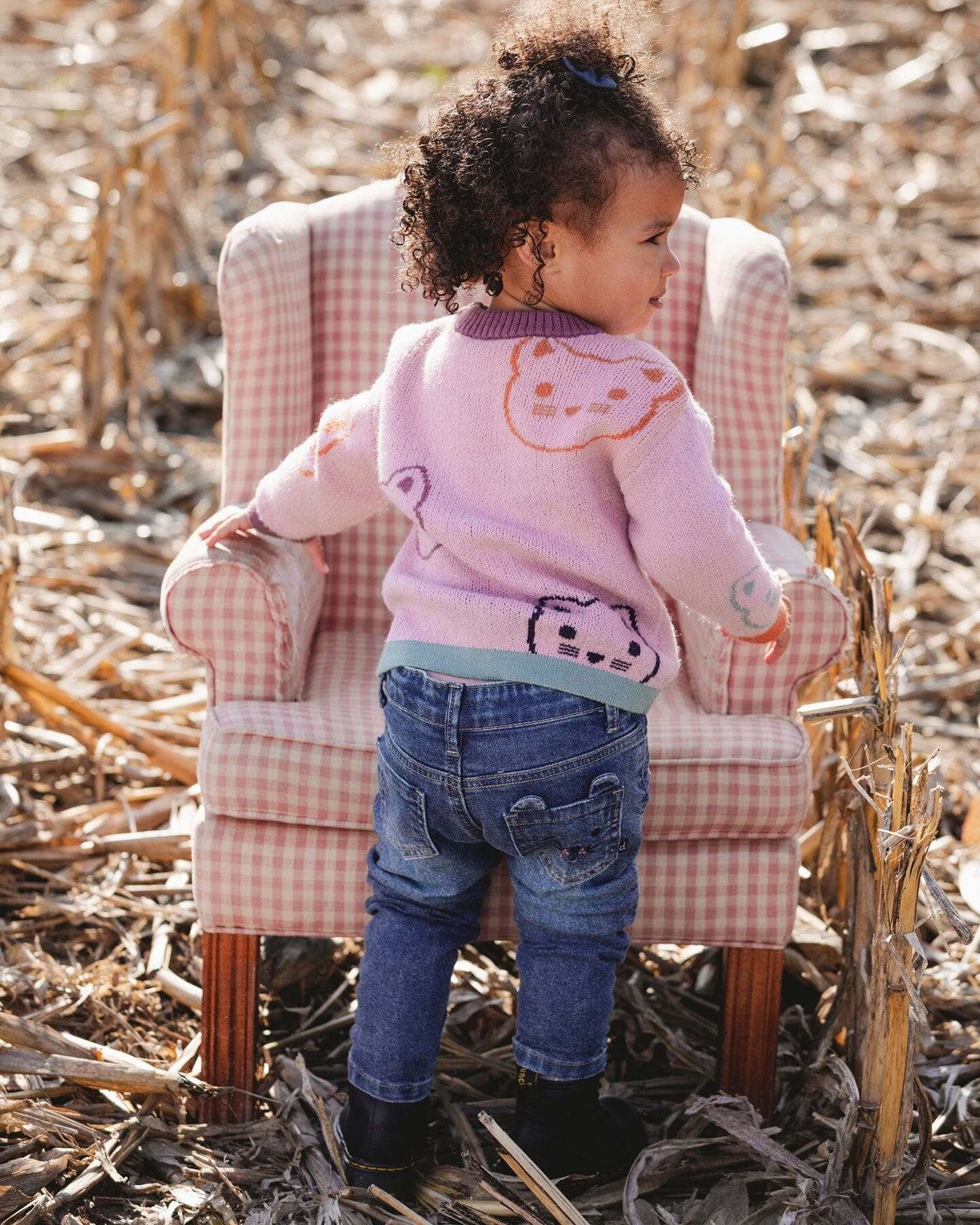Toddler wearing blue denim jeans with embroidered details, standing in a field next to a pink gingham armchair.
