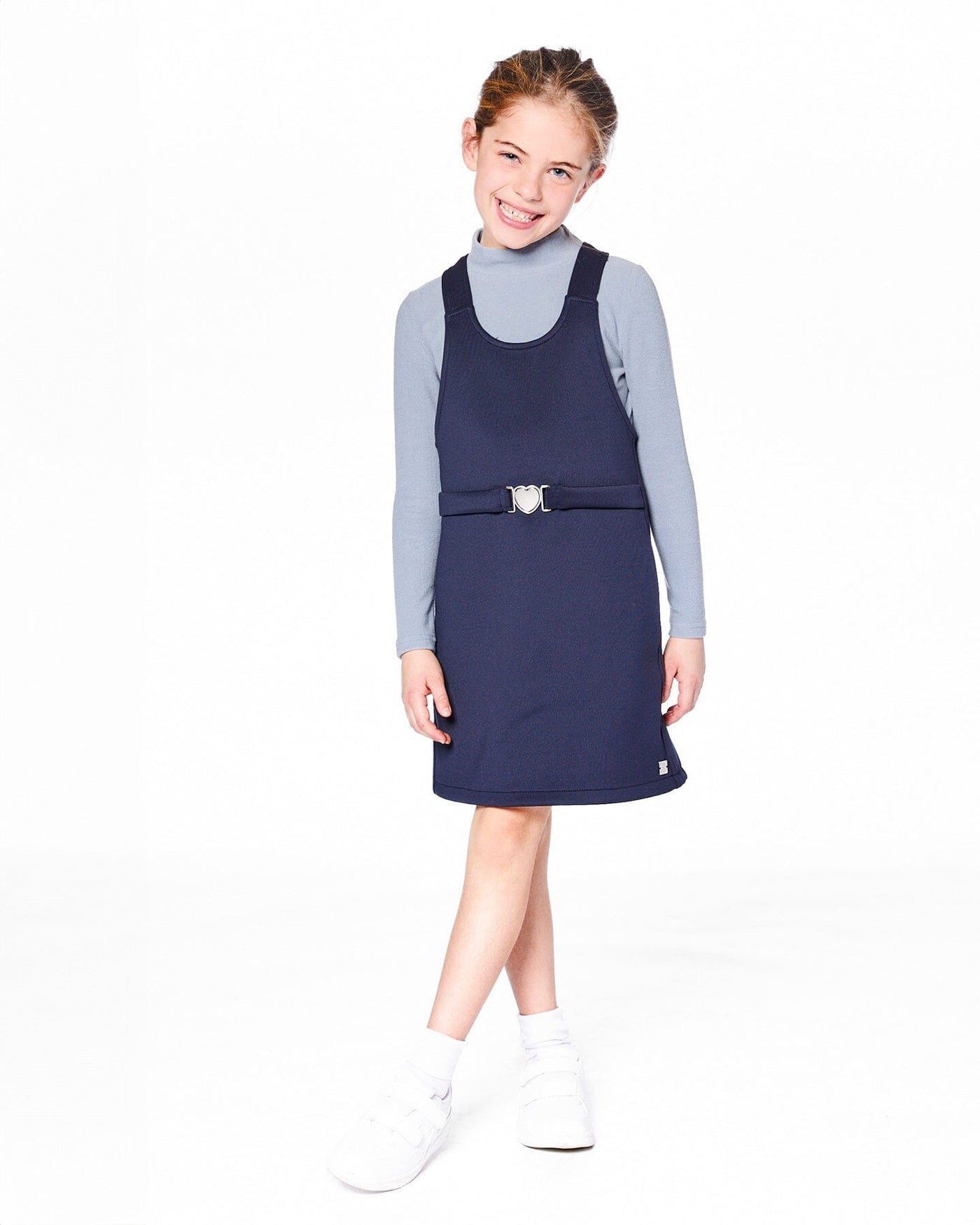 Young girl wearing navy blue Milano jumper dress with wide straps and silver buckle detail over a light blue long-sleeve top