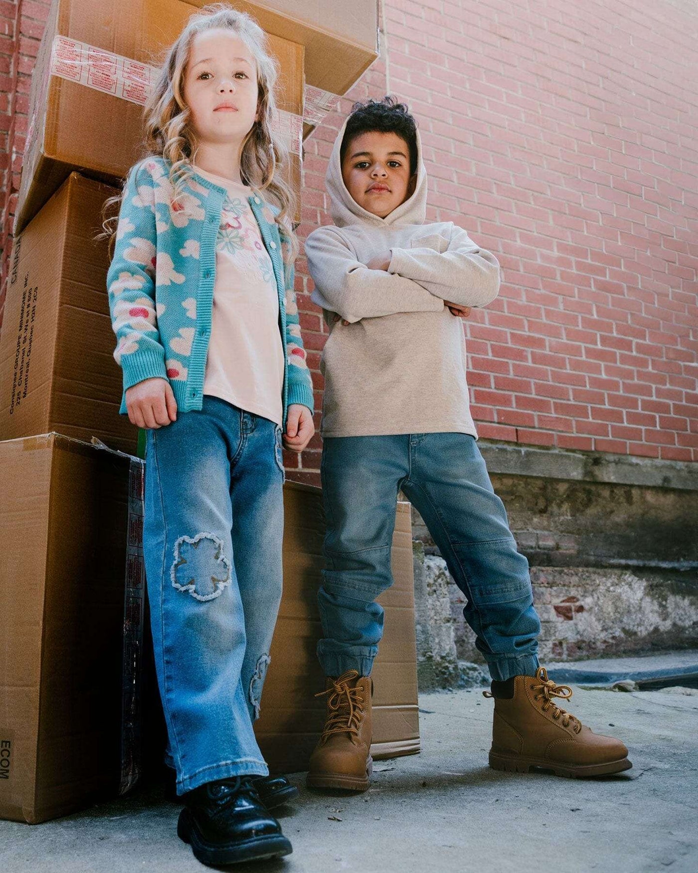 Two kids wearing casual outfits including light blue denim French Terry jogger pants and boots standing outdoors near stacked cardboard boxes and a brick wall
