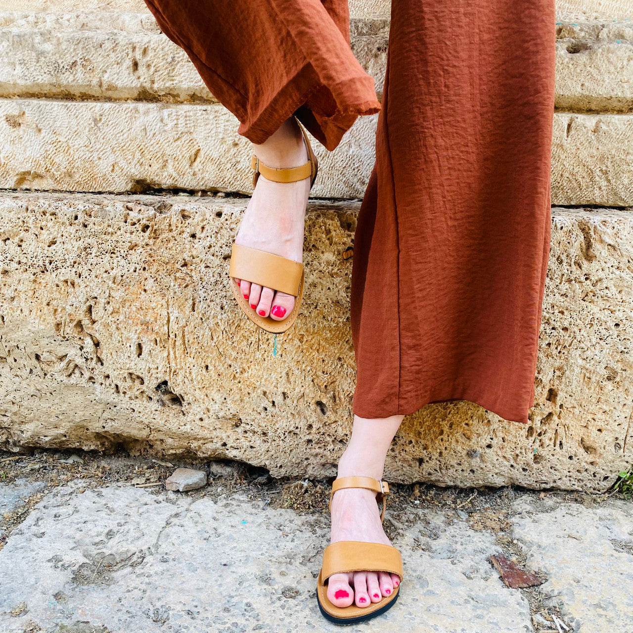 Close-up of feet wearing tan Aventura Leather Walking Sandals with ankle straps, standing on stone steps, paired with rust-colored pants