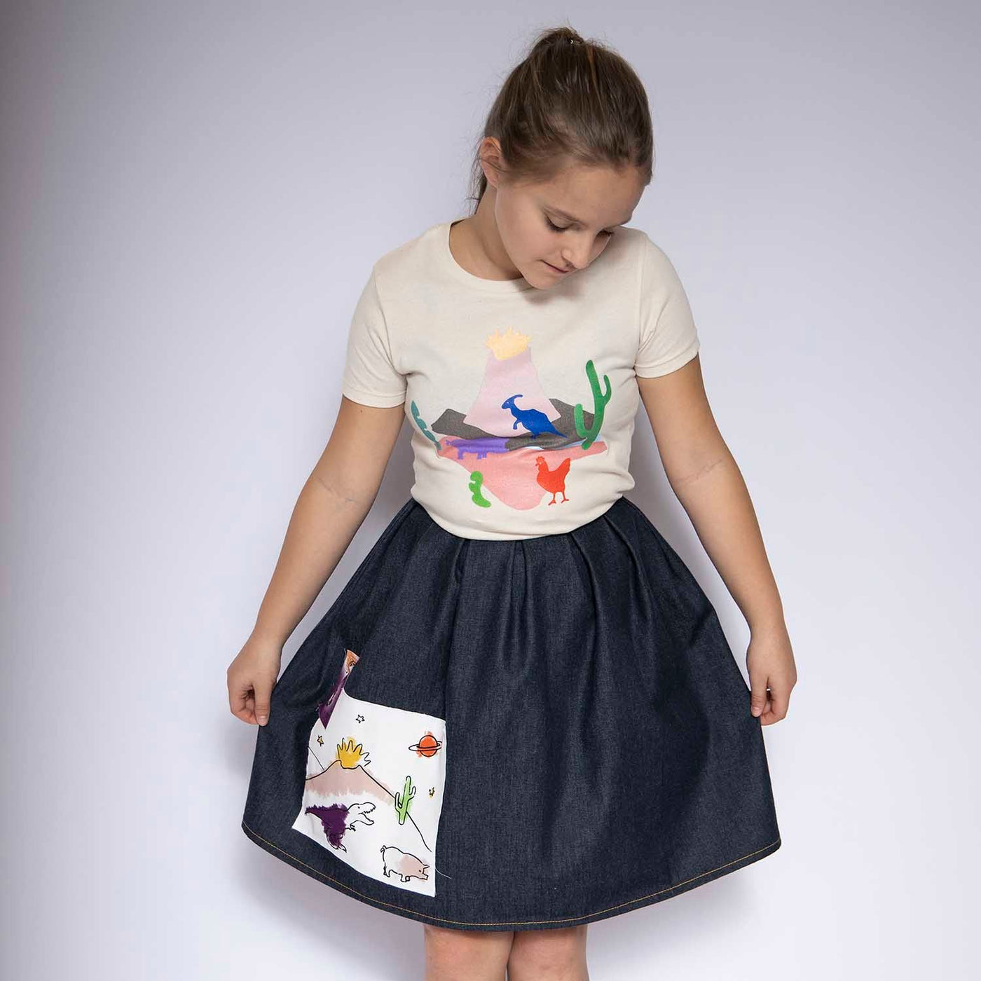 Girl wearing a blue denim skirt with colorful appliqué and a printed t-shirt against a plain background