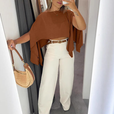 Woman wearing solid color off-shoulder brown top with bell sleeves and white high-waist pants, holding a woven handbag in a minimalist room
