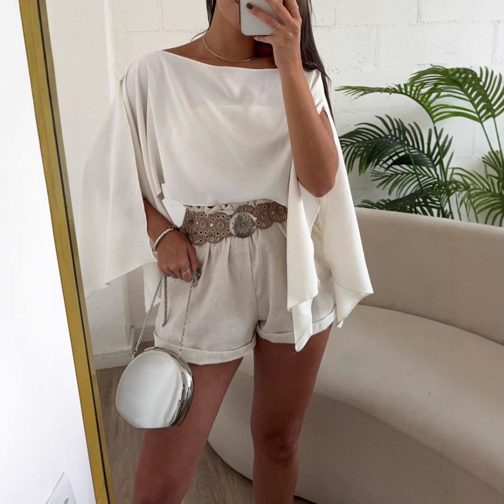 Woman wearing white off-shoulder top with bell sleeves and beige shorts, posing indoors with a round silver handbag
