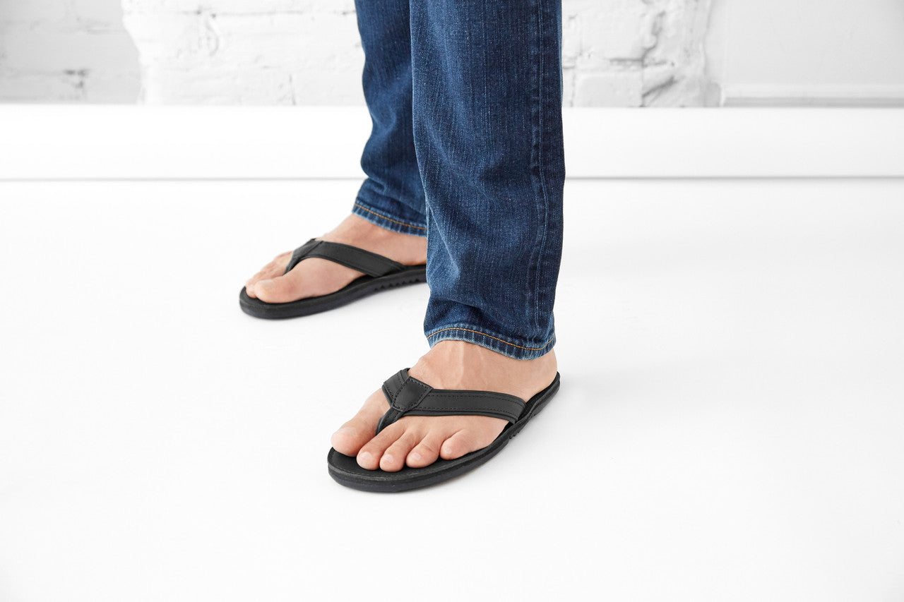 The Padre men's black leather flip flops with cushioned sole and durable tire tread, worn with blue jeans on white floor