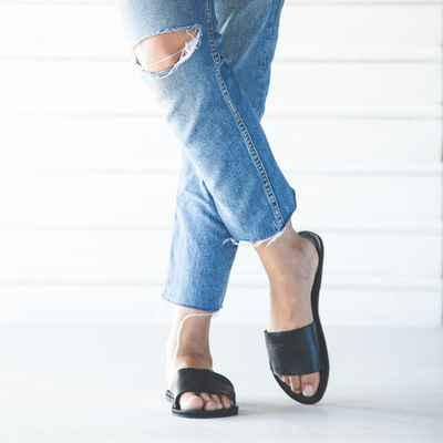 The Linda Leather Slide black leather open-toe sandal worn with ripped blue jeans on a white background