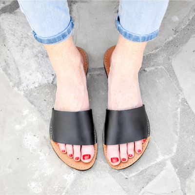 The Linda Leather Slide sandal in black leather with hand-cut sole and cushy mid-sole worn with rolled light blue jeans on stone floor