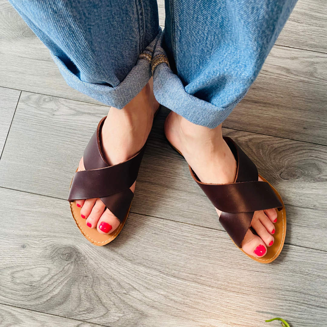 Woman wearing genuine leather slides with crossed brown straps and red nail polish on wooden floor