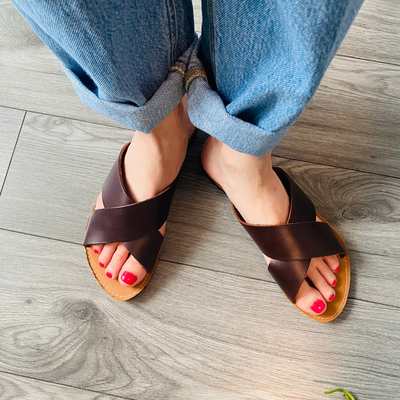 Woman wearing genuine leather slides with crossed brown straps and red nail polish on wooden floor