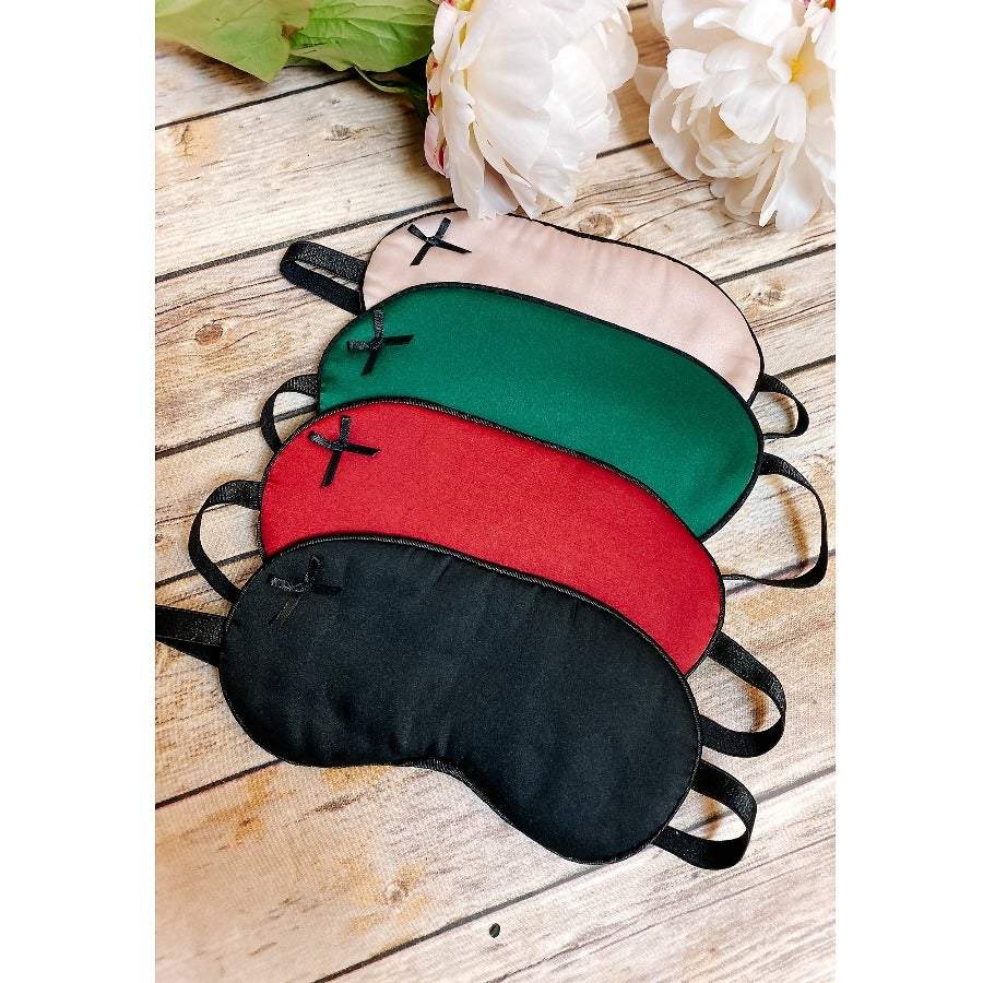 Set of four satin sleep masks in black, red, green, and pink colors with small bow accents on wooden surface with white flowers.