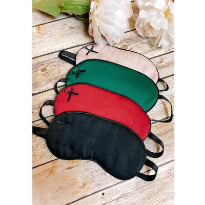 Set of four satin sleep masks in black, red, green, and pink colors with small bow accents on wooden surface with white flowers.