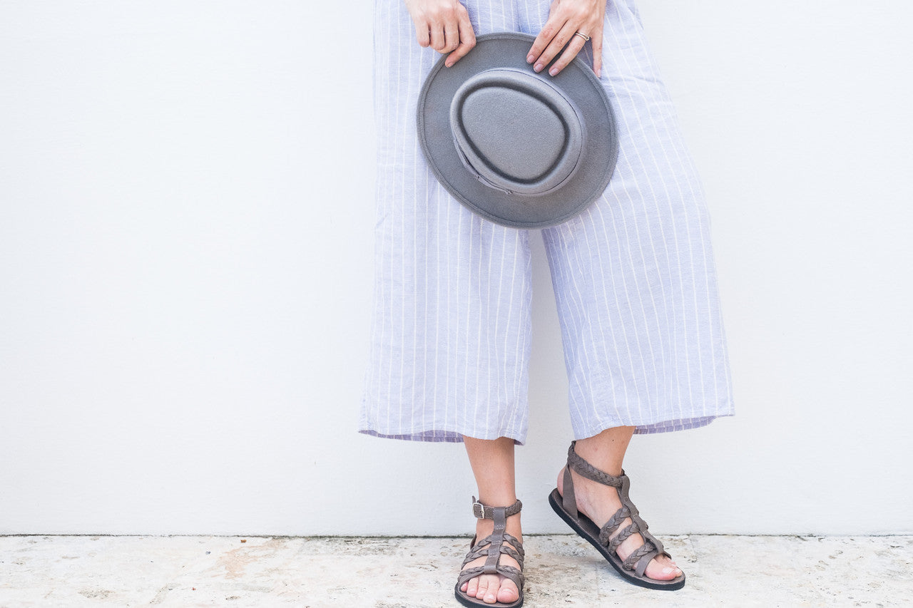 The Kaitlin Gladiator Leather Sandal worn with light striped pants and holding a gray hat, featuring handcrafted leather straps and upcycled tire soles.